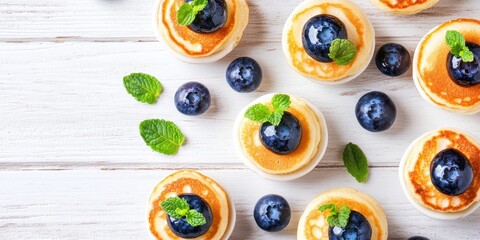 Fluffy mini pancakes with blueberries and mint on a white wooden backdrop arranged aerially for a fresh healthy food concept