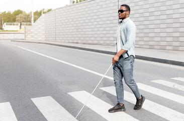 Young black visually impaired man wearing dark glasses, walking across city street, using cane....