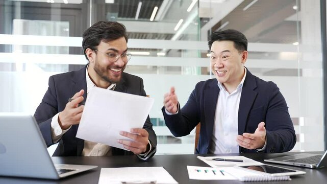 Two excited businessmen celebrate successful results in office setting. They show joy and excitement while reviewing documents and charts. Teamwork and accomplishment in a professional environment