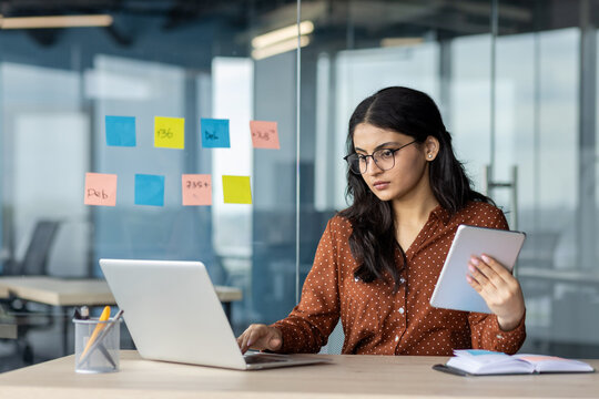 Focused but serious businesswoman working with laptop inside office. Woman holding tablet computer and typing on keyboard. Office worker at workplace confident and successful.