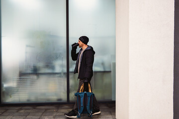 A man with a backpack walks against the background of modern urban architecture. He has a cup with a warm drink in his hands. He is dressed in a black autumn jacket and jeans.