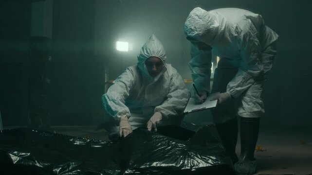 Full lowkey shot of young Caucasian female forensic expert wearing protective overalls examining dead body under black bag with male colleague writing down report under blinking police light