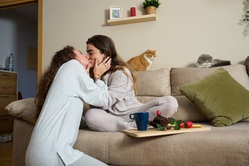 Lesbian couple kissing on sofa with cats and valentine's day breakfast