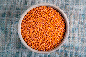 A wooden bowl with red lentils grains on a burlap. Top view of dried orange lentils inside the bowl.