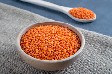 Red lentil grains in a wooden bowl and spoon on a burlap. Top view of dried orange lentils inside the bowl.