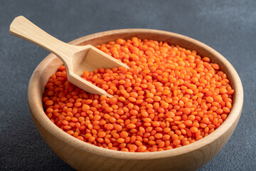 Red lentil grains in a wooden bowl and spoon on a gray background. Top view of dried orange lentils inside the bowl.