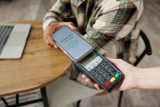 Cropped young man in casual clothes hold wireless bank payment terminal process acquire blank screen mobile cell phone sitting at table in coffee shop cafe relax rest in restaurant during free time.