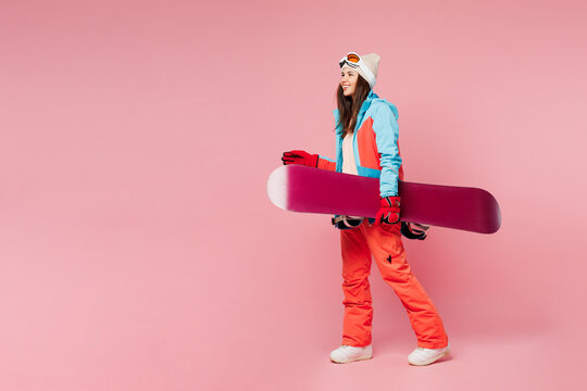 Full body side view young smiling woman wear warm blue padded jacket ski goggles mask spend weekend winter season in mountains hold snowboard walk go isolated on plain pink background. Hobby concept.