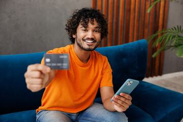 Young man wear casual clothes sits on blue sofa couch use mobile cell phone give credit bank card shopping online stay home hotel flat rest relax spend free time in living room indoor. Lounge concept.