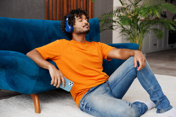 Young man wearing casual clothes sits near blue sofa couch use mobile cell phone listen music in...