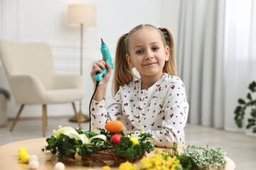 Little girl with hot glue gun creating Easter composition at table indoors