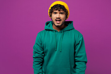 Young sad shocked mad indignant dissatisfied displeased Indian man he wear green hoody yellow hat casual clothes looking camera isolated on plain purple background studio portrait. Lifestyle concept.