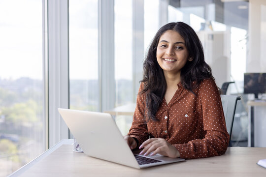 Portrait young successful woman at her workplace inside the office. Businesswoman smiling and looking at camera, typing on the laptop keyboard, sitting by the window, happy and satisfied employee