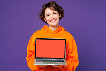 Naklejka premium Young smiling happy Caucasian IT woman she wear orange hoody casual clothes hold use work on laptop pc computer with blank screen workspace area isolated on plain purple background. Lifestyle concept.
