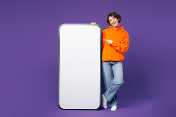 Full body young woman she wears orange hoody casual clothes point index finger on big huge blank screen mobile cell phone smartphone with area isolated on plain purple background. Lifestyle concept.