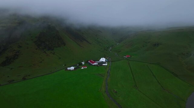 A drone flies through the clouds over lush meadows and mountains in Iceland, passing above houses nestled near a waterfall, capturing the serene and majestic landscape.