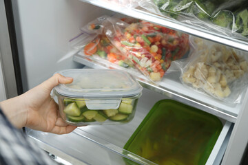 Woman taking glass container with frozen chopped zucchini from refrigerator, closeup