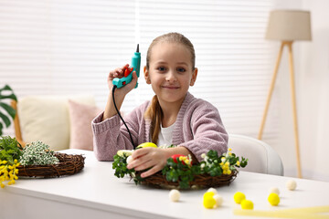 Little girl with hot glue gun creating Easter composition at table indoors