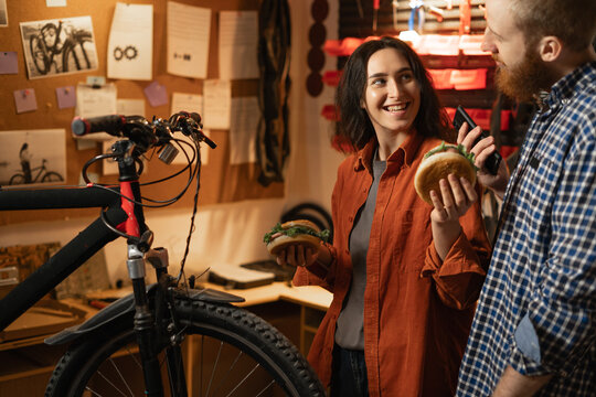 Young couple or friends eating burgers working together in professional bike repair workshop - Powered by Adobe