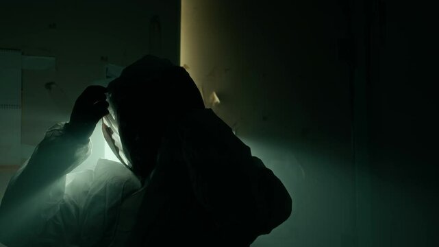 Medium lowkey slowmo portrait of forensic expert finishing work at crime scene taking off protective hood and cap, illuminated by backlight in dark grim building