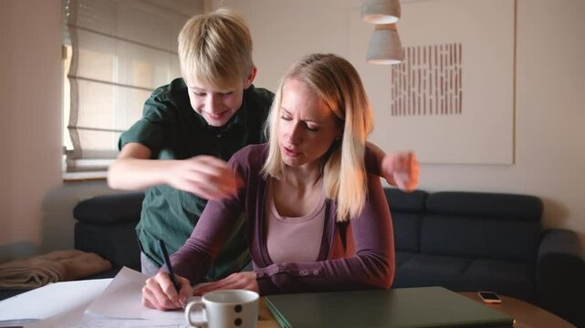 
A mother is working from home on her laptop in the living room when her son comes up and embraces her. She pauses her work to share a loving hug with him.