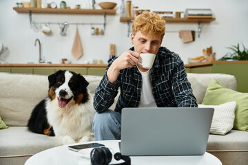 A young redhead man enjoys a warm drink while working on a laptop at home with his dog.
