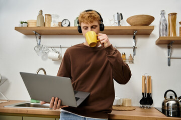 Young man with red hair focuses on his laptop while sipping coffee in a chic kitchen.