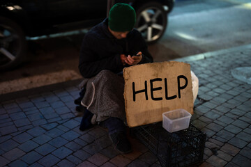 A homeless man is outside in the winter, with a cardboard sign in front of him that says 