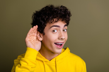 Cheerful young man in yellow hoodie listening attentively with a hand gesture against a khaki...