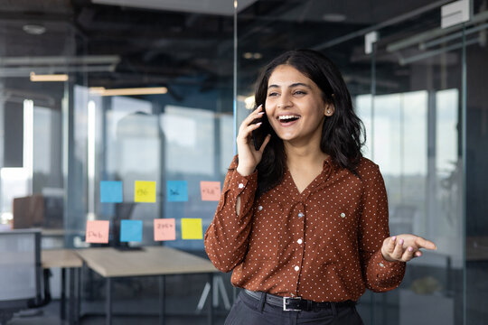 Successful and happy office worker talking on the phone while standing near the window inside the office building. Satisfied businesswoman at workplace communicating with colleagues and clients.