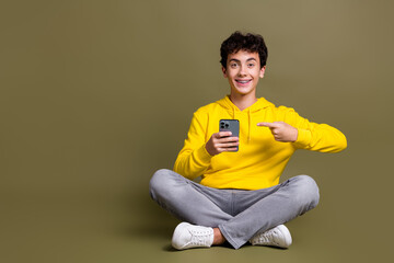 Teenager in yellow hoodie smiling and sitting cross-legged while using a smartphone against a khaki...
