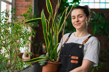 Running of own business. Young woman florist holding plant in pot wearing apron in botanical store....