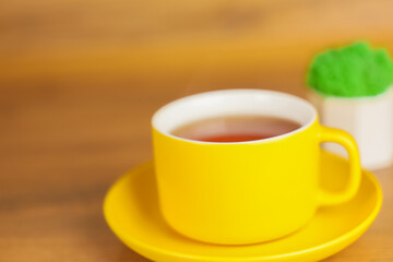 Side view of a yellow cup with saucer in which black tea is poured stands on a wooden table on the left with space for text, next to it is a plant in a white pot. Concept of tea drinking, cleaning