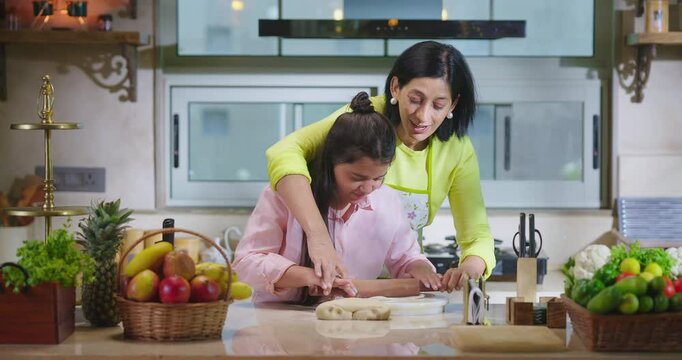 Indian happy woman lady with cute teen age cute girl rolling raw flour dough learn cooking food indoor home smiling young mom teach to kid child baking cookies enjoy day indoor modern home kitchen
