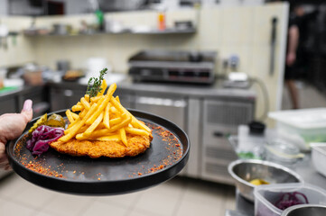 A close-up of a hand holding a black plate with golden fries, chicken schnitzel, pickled vegetables, and a garnish of thyme, set in a bustling restaurant kitchen background.
