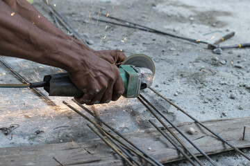 The brown skinned hands of a male craftsman hold a grinder in his right hand, and his left hand...
