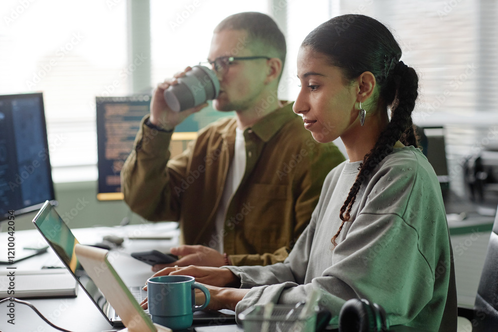 Poster side view of smiling middle eastern female programmer typing at ease on laptop sitting at cluttered 