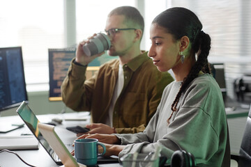 Side view of smiling Middle Eastern female programmer typing at ease on laptop sitting at cluttered...
