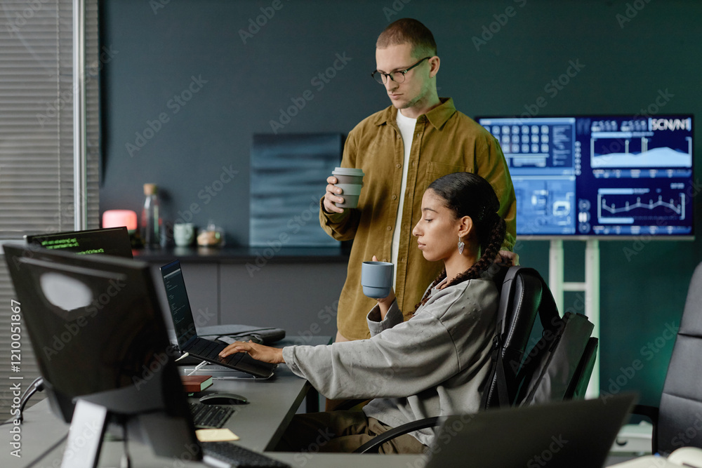 Poster side view of female it programmer typing on laptop collaborating with male colleague over cup of ref