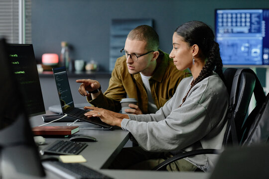 Side view of male IT programmer pointing at laptop screen helping female colleague with code debugging while collaborating at corporate workplace
