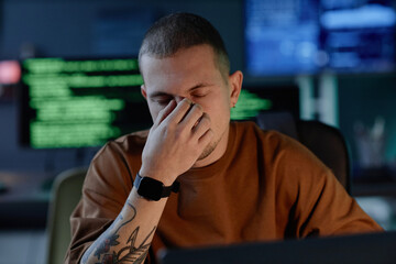 Portrait shot of male programmer exhausted from work overload sitting at desk with eyes closed...