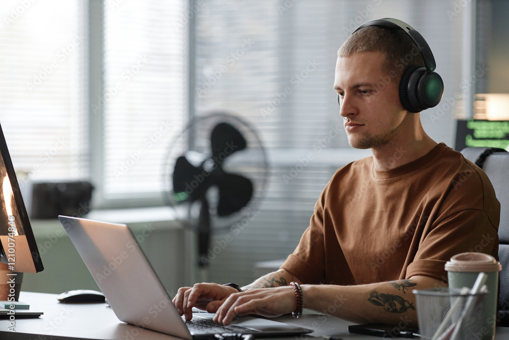 Poster medium shot of handsome male programmer wearing headphones coding on laptop while working at desk in