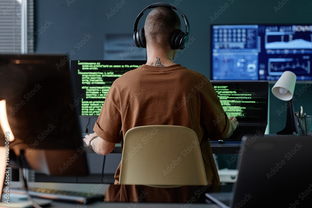Poster rear view of male it developer wearing headphones immersed in writing code on multiple computers at 