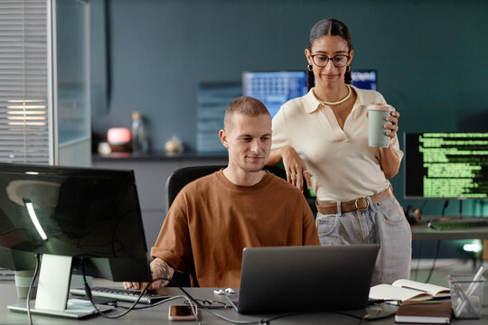 Middle Eastern female senior developer assisting cheerful male junior specialist looking at code on laptop screen during working meeting in office