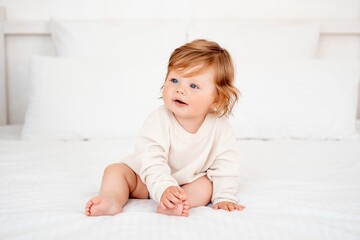 A baby with red hair and blue eyes in a white bodysuit is smiling happily on the bed at home, a healthy happy cute newborn baby boy