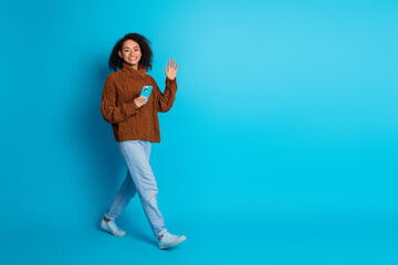 Young woman holding a phone and waving against a blue background, smiling and wearing a brown...