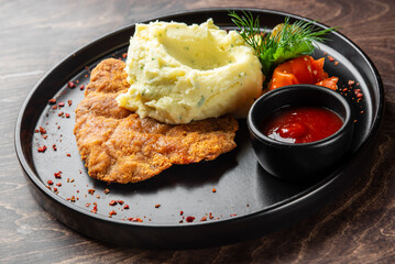 A delicious serving of crispy fried chicken with creamy mashed potatoes, accompanied by a side of mixed vegetables, pickles, and a small bowl of ketchup, all on a black plate.