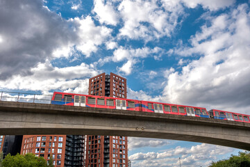 A vibrant red train travels over a concrete bridge, set against a backdrop of dramatic clouds and modern architecture, embodying urban movement and connectivity in London UK