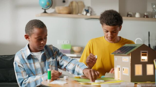 Two brothers work on school science project in cozy room. Younger assembles with glue, older cuts paper. Solar-powered cardboard house model glows nearby. Concept of teamwork and renewable energy