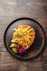 A delicious plate featuring a crispy breaded chicken filet topped with golden french fries, garnished with fresh thyme, and accompanied by pickles and pickled onions on a rustic wooden table.